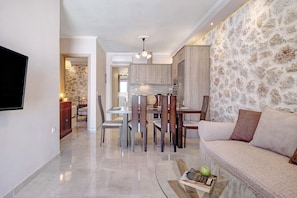 A modern living room with a sofa, a dining area with chairs, a kitchen with cabinets, and a TV mounted on the wall.