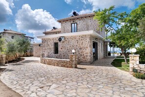 Edifici de pedra amb teulada a mànega, entrada pavimentada i mur de pedra amb placa.