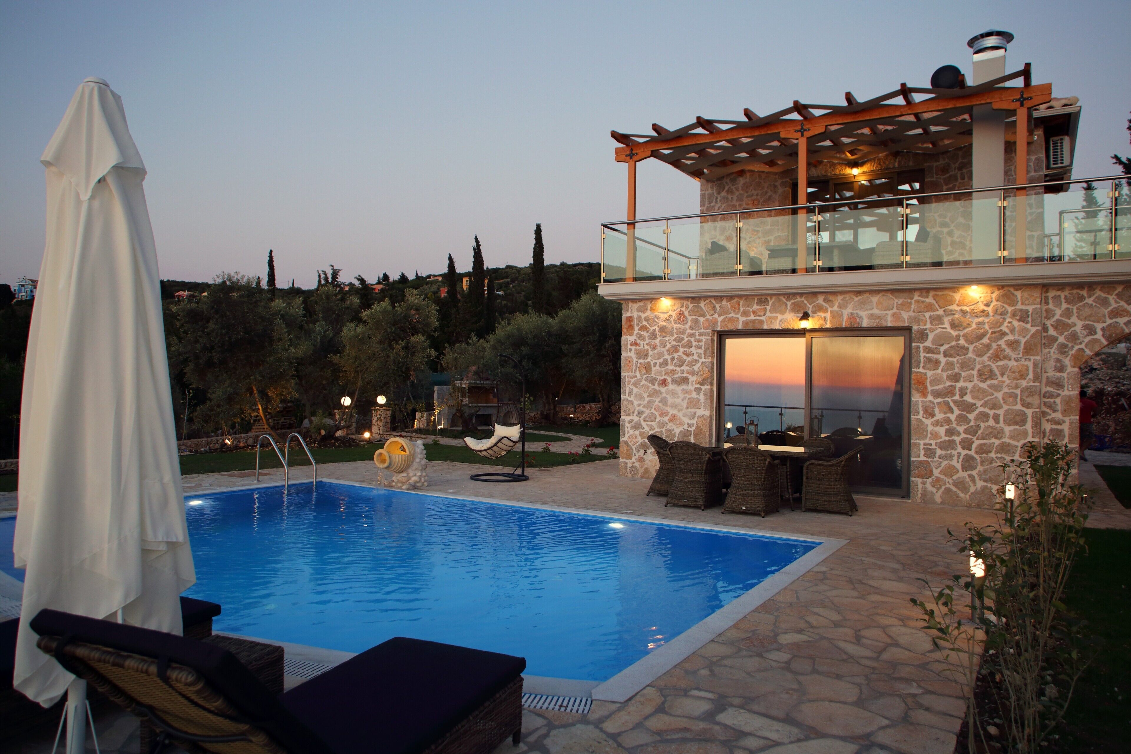 Une piscine avec des chaises longues, un patio couvert et un bâtiment en pierre doté d’un balcon en verre.