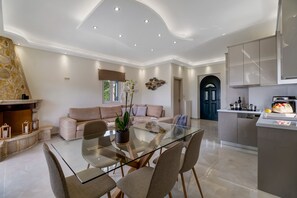 Una sala de estar moderna con una mesa de comedor de vidrio, sofás color beige, una chimenea y una zona de cocina con armarios y electrodomésticos.