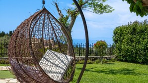 Una silla colgante con cojín, un patio de piedra, una valla metálica y vista al mar.