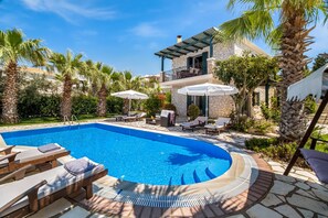 A swimming pool with lounge chairs and umbrellas, surrounded by palm trees and a stone building.