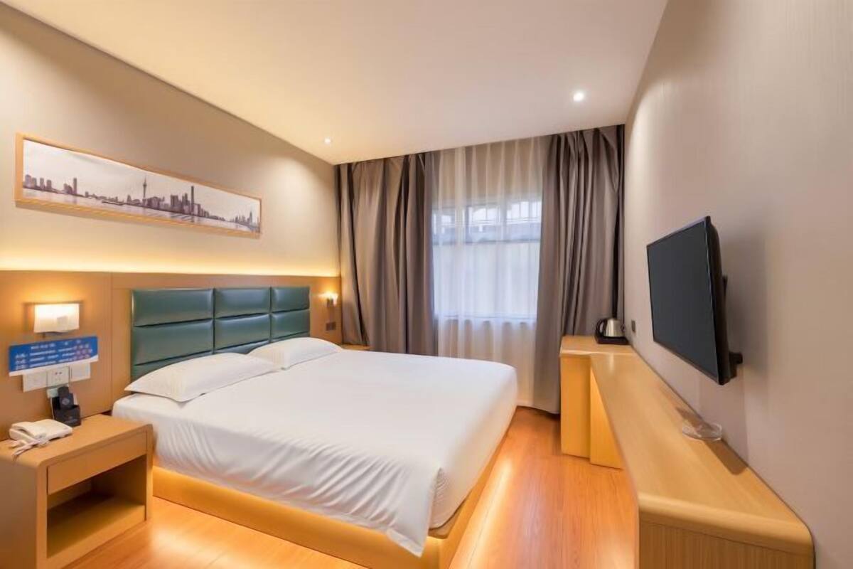 A hotel room with a bed, bedside table, lamp, TV, and a cityscape painting on the wall.