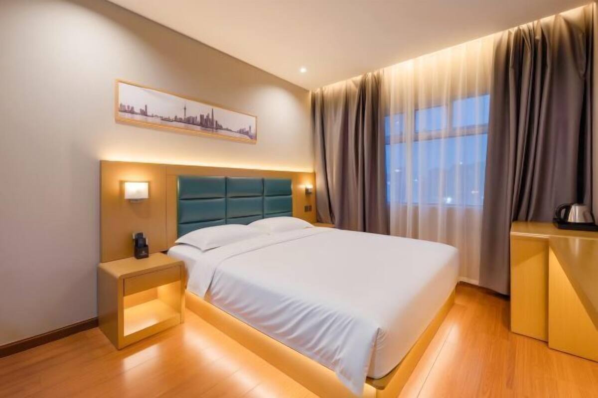 A hotel room with a bed, bedside table, lamp, and a cityscape painting on the wall.