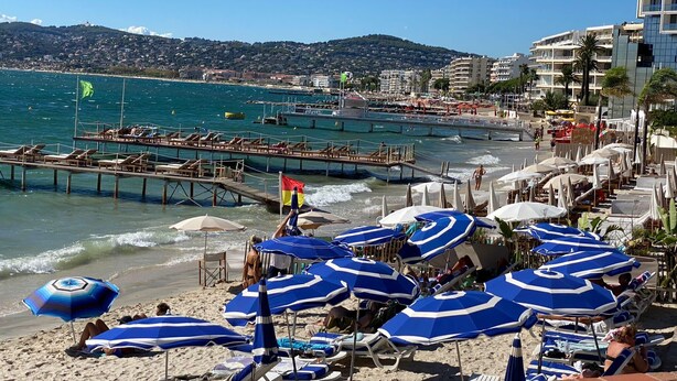 Una playa con sombrillas azules, sillas de descanso y un muelle que se adentra en el agua.
