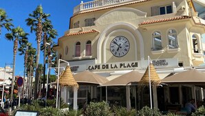 Un edificio de varios pisos con un café llamado "Cafe de la Plage", con terraza y un reloj grande.