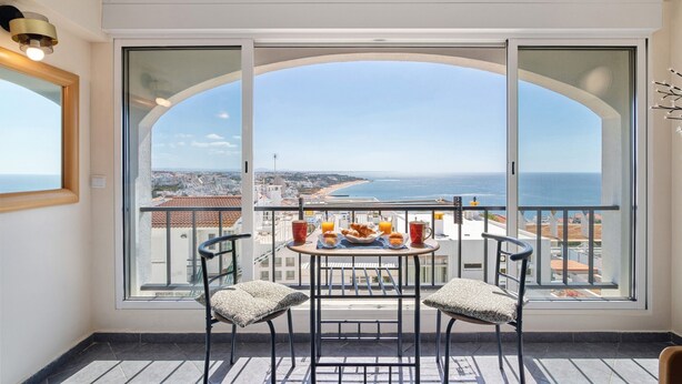 A balcony with a table set for breakfast, two chairs, and a large window offering a view of the sea and cityscape.