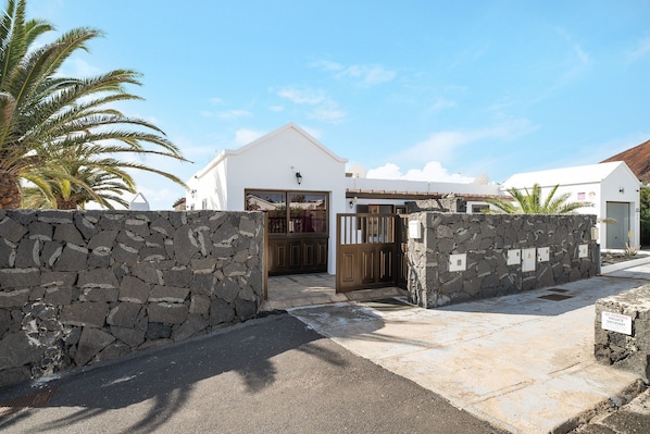 A stone wall entrance to a building with a white facade, a wooden gate, and a palm tree in the background.