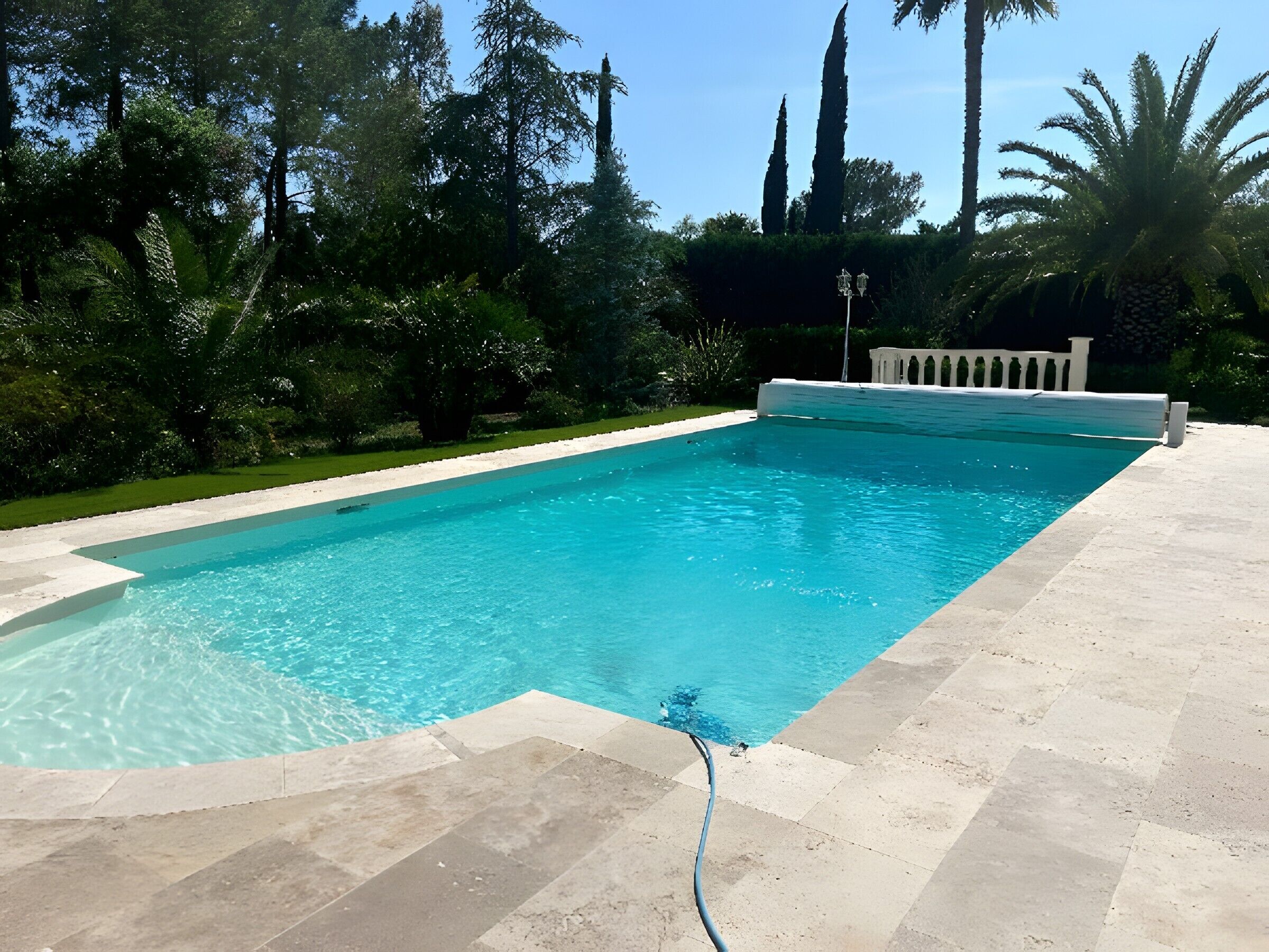 A rectangular swimming pool with clear blue water, surrounded by a tiled deck and lush greenery.