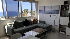 Ein modernes Wohnzimmer mit grauem Sofa, einem Couchtisch, einer Stehlampe und einem Bücherregal. Ein großes Fenster bietet Blick auf das Meer und Palmen.