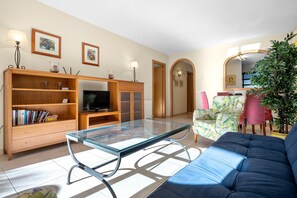 Un salon comprenant une table basse avec un plateau en verre, un canapé bleu, un meuble de rangement en bois avec un téléviseur et une bibliothèque.