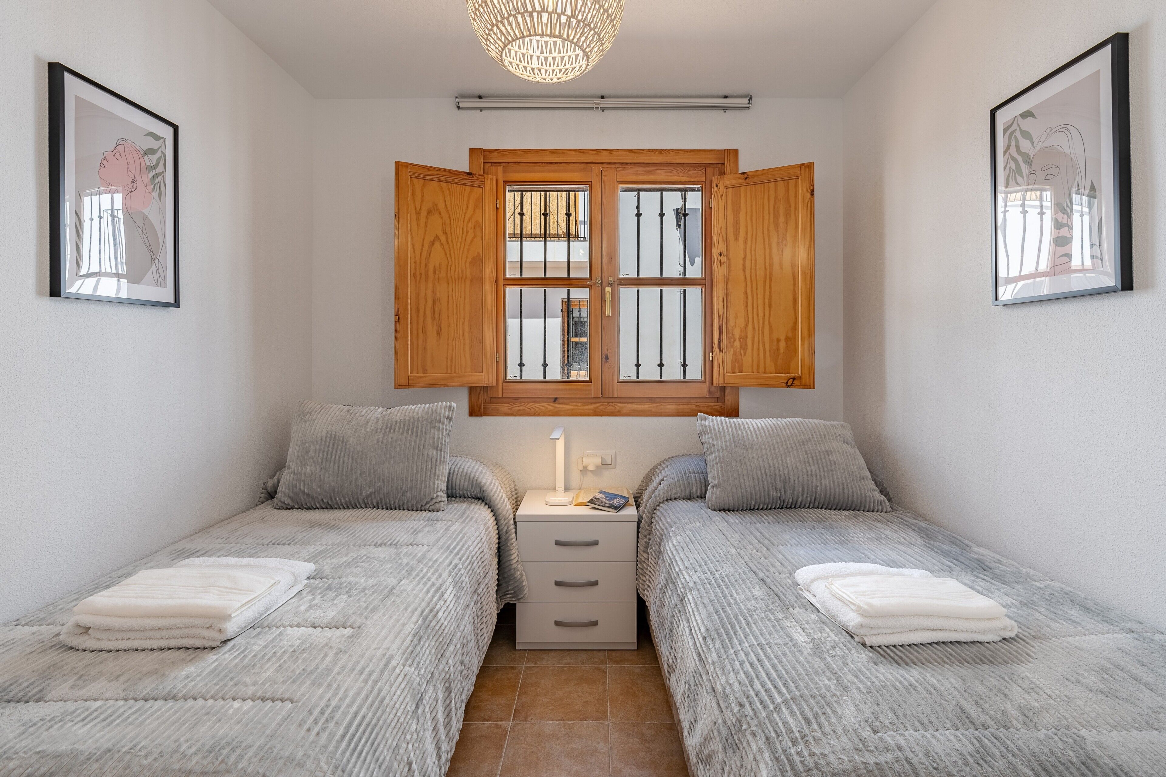 A bedroom with two beds, a nightstand, a window with wooden shutters, and framed artwork on the walls.