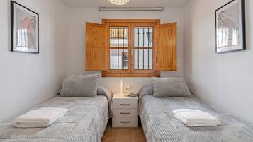 A bedroom with two beds, a nightstand, a window with wooden shutters, and framed artwork on the walls.