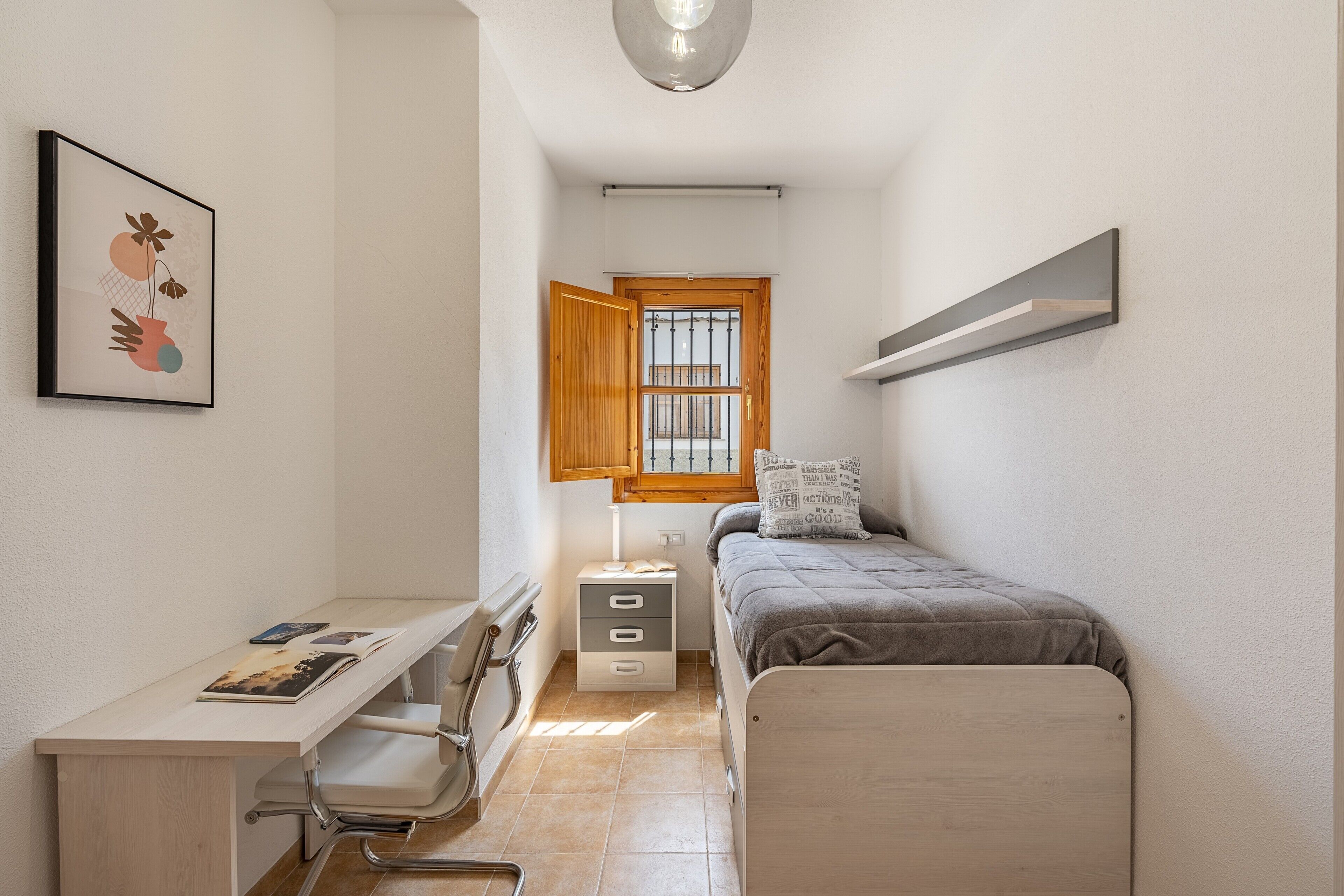 A bedroom with a bed, desk, chair, bedside table, and a window with a wooden blind.
