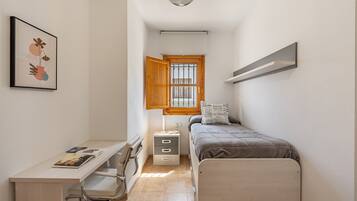 A bedroom with a bed, desk, chair, bedside table, and a window with a wooden blind.