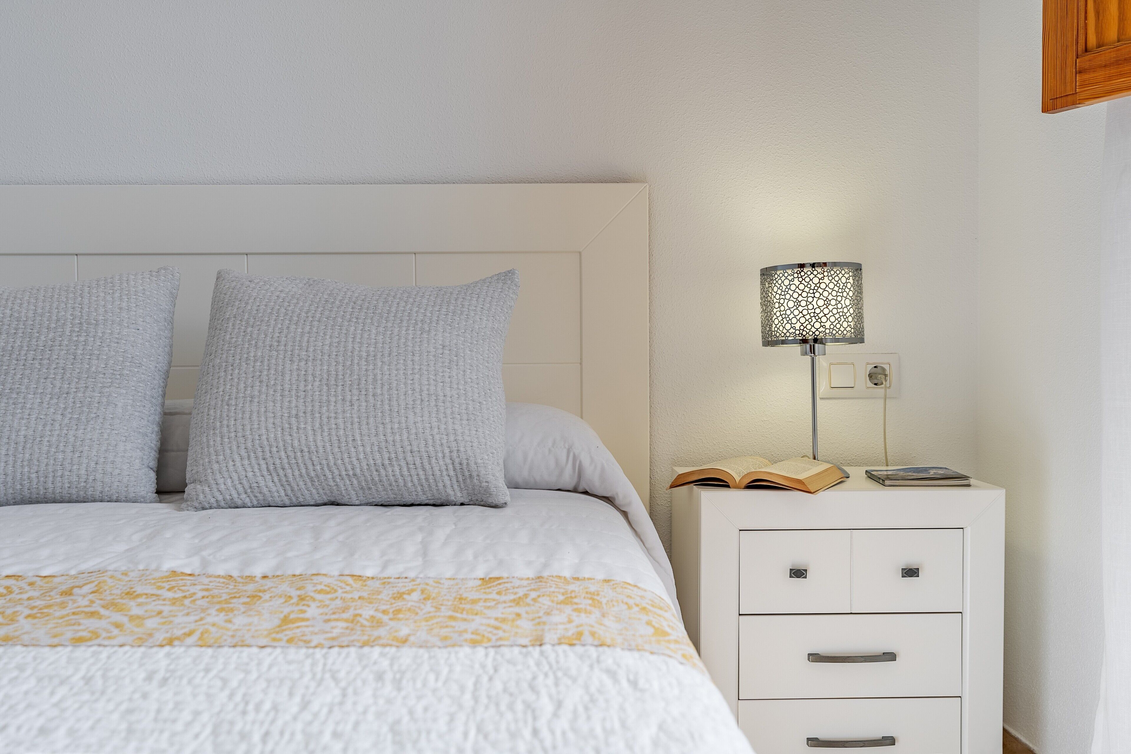 Una cama bien hecha con una colcha estampada, dos almohadas, una lámpara de noche y una mesita de noche con un libro y una revista.