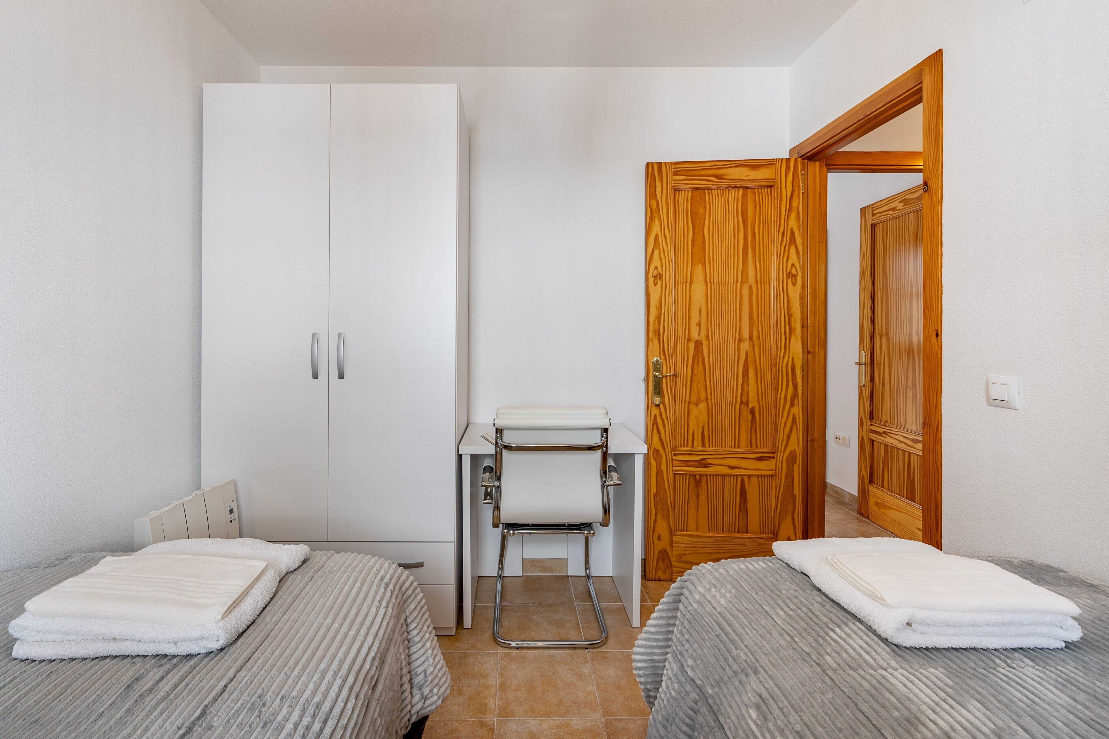 A small room with two beds, a white wardrobe, a wooden door, and a small table with a chair.