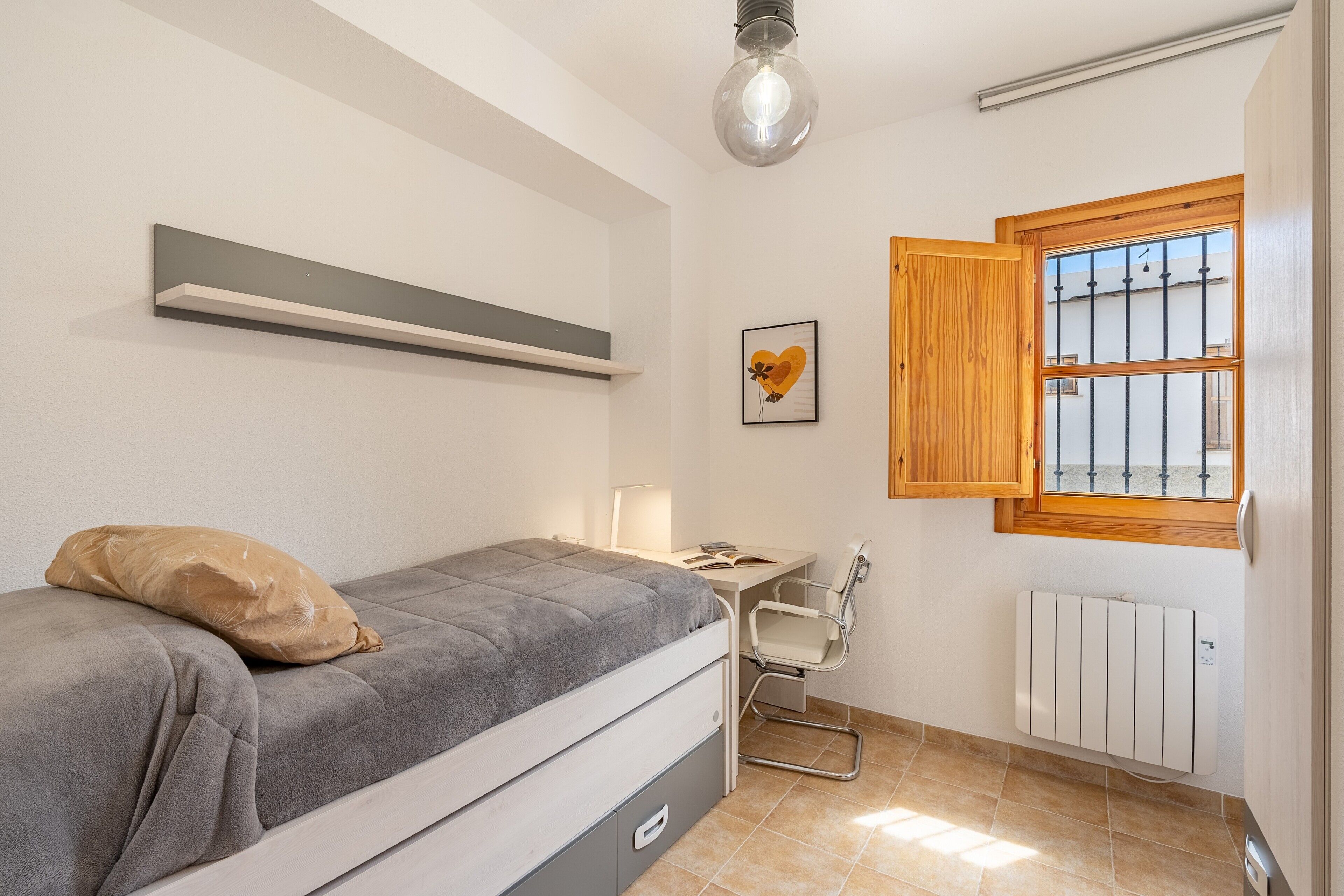 Un dormitorio con cama, escritorio, silla, radiador, ventana con contraventanas de madera y un cuadro en la pared.