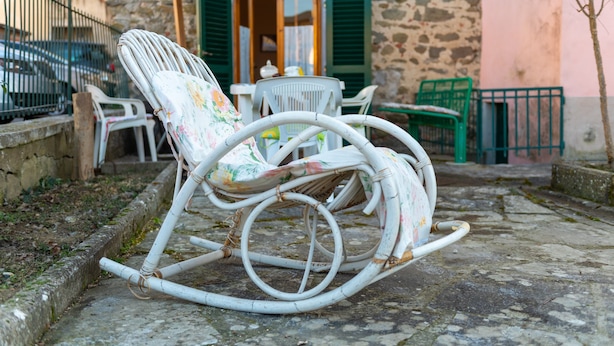 Un espacio de estar al aire libre rústico con una mecedora de mimbre, sillas de plástico blanco y una mesa con tetera.