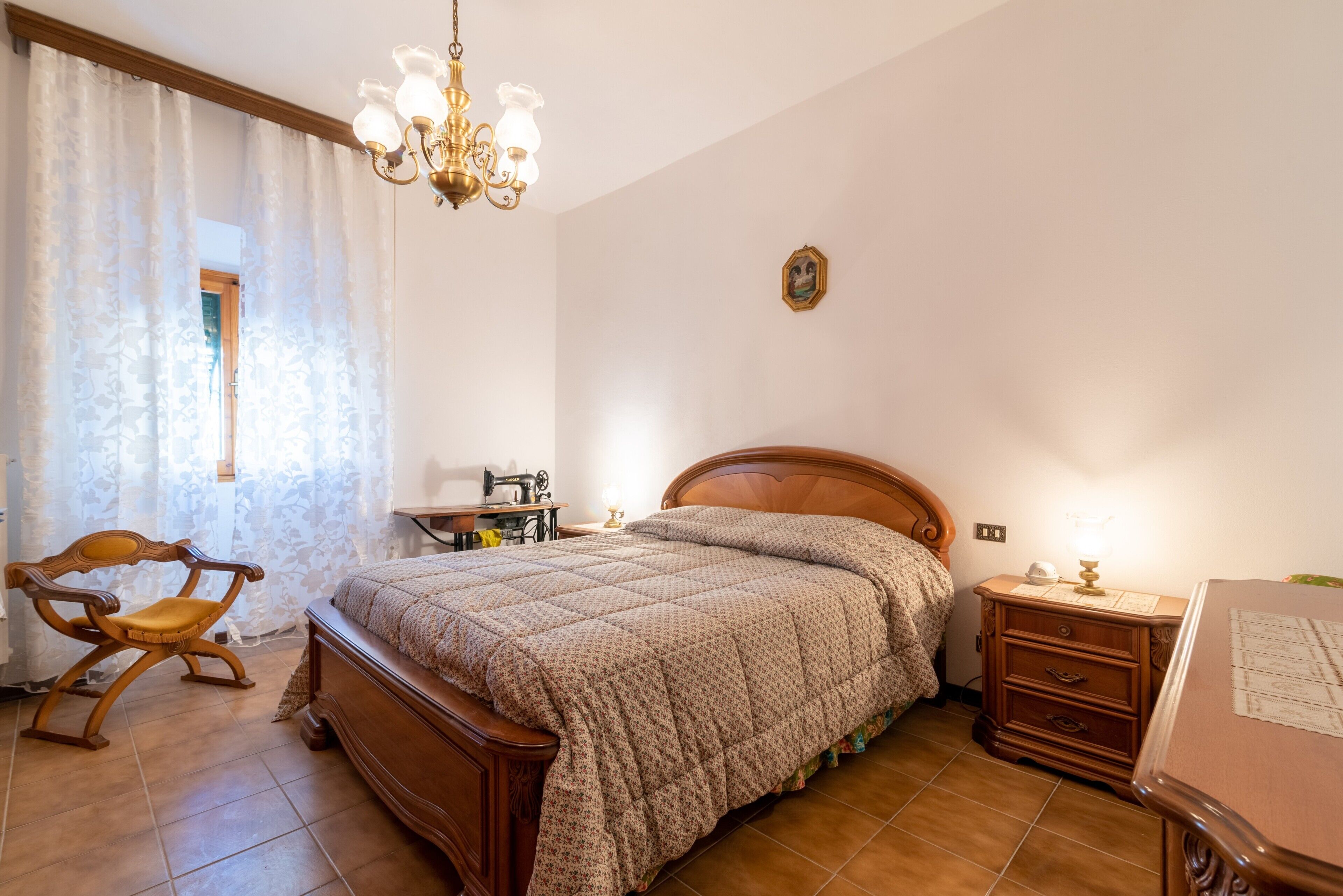 A bedroom with a wooden bed, a nightstand, a chair, a sewing machine, and a chandelier.