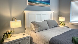 A bedroom with a bed, nightstands, a wall-mounted lamp, a window with blinds, and a painting of a beach scene. - Welcome to The 255 — Your Home Away from Home (Parkland)