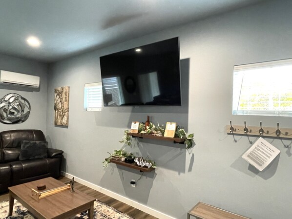 Un salon avec un téléviseur à écran plat fixé au mur, un canapé en cuir, une table basse et une étagère ornée de plantes et d’une pancarte de bienvenue.