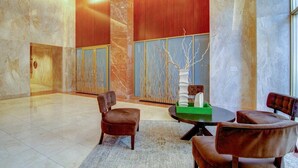 Eine geräumige Lobby mit Marmorwänden, einer großen Holzverkleidung und einer dekorativen Skulptur von einem Baum. Es sind Lederstühle und ein runder Tisch mit einer grünen Ablage vorhanden.
