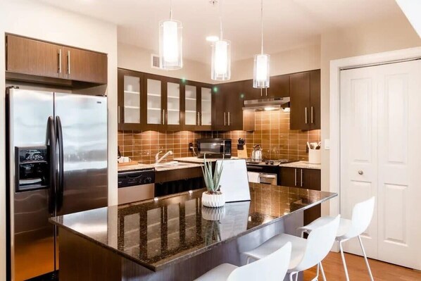 A modern kitchen with a stainless steel refrigerator, dark wooden cabinets, a granite countertop island, and white chairs.