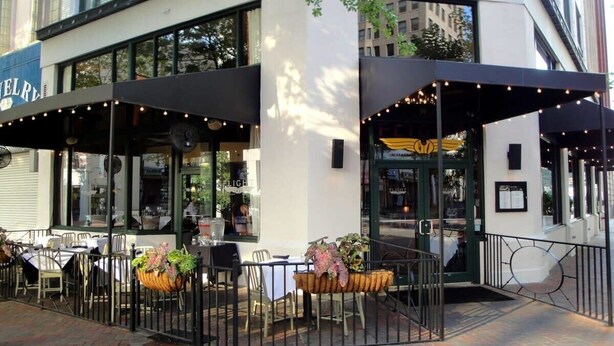 Un área de comedor al aire libre con mesas y sillas, una zona de estar cubierta y la entrada de un edificio con puerta y ventanas.