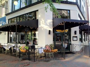 An outdoor dining area with tables and chairs, a covered seating area, and a building entrance with a door and windows.