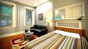 Une chambre d’hôtel avec un lit, un canapé, une petite table et deux fenêtres équipées de stores.
