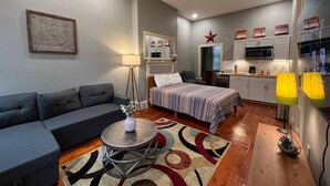 Un espace de vie compact comprenant un canapé, un lit avec une couvre-lit à rayures, une petite table ronde avec un vase, et un coin cuisine avec des placards et un four à micro-ondes.