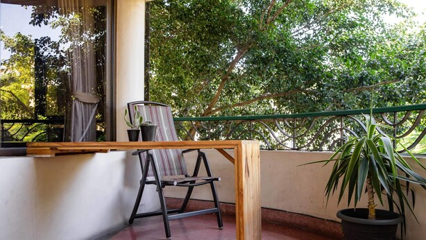 Ein Balkon mit Holztisch, einem Stuhl, einem Blumentopf und Blick auf Bäume.