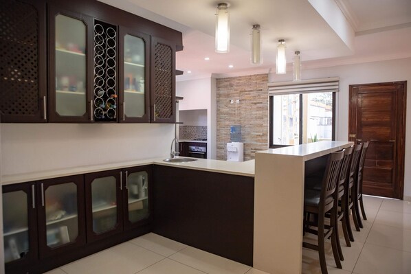 A modern kitchen with dark cabinetry, a central island with bar stools, and a large window providing natural light. - Maison Moz - Sommerschield III (Maputo)