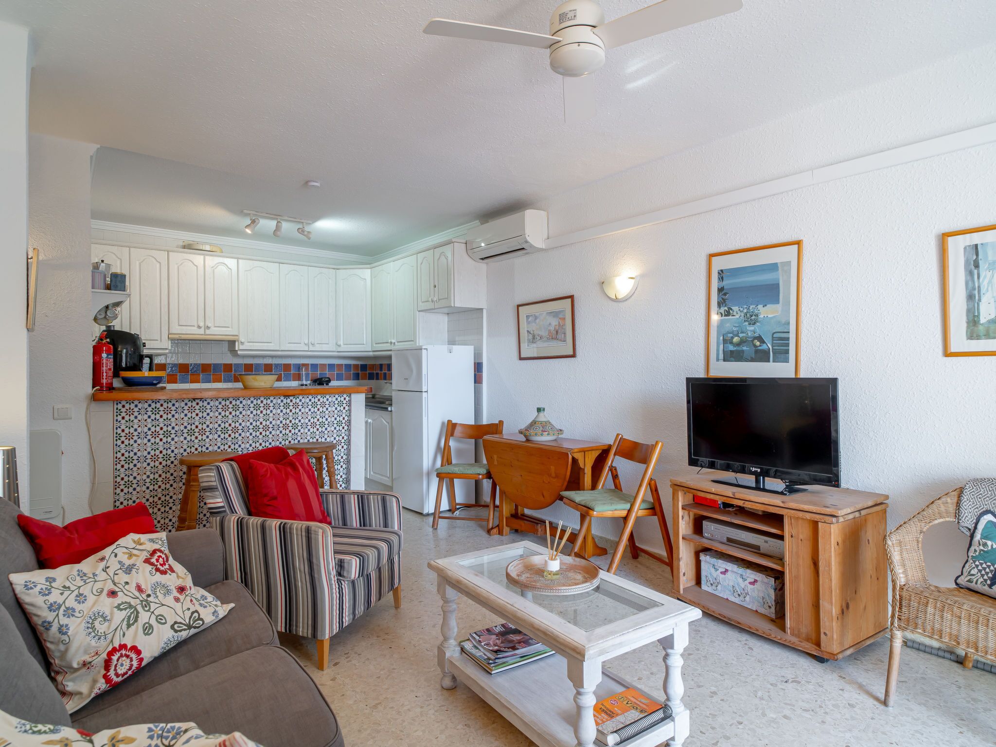 Una sala de estar con sofá, sillón, mesa de centro y televisor. Cuenta con una zona de cocina con armarios y una mesa de comedor con sillas.