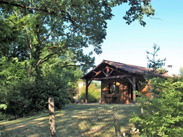 A rustic wooden cabin with a covered patio, surrounded by dense foliage and a chain-link fence.