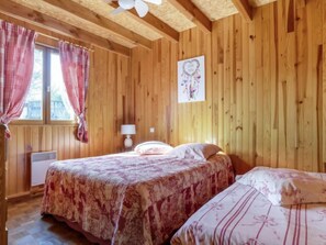 A wooden cabin bedroom with two beds, a ceiling fan, a window with curtains, and a wall-mounted lamp.