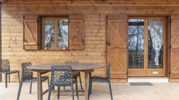 Une cabane en bois avec une table ronde et des chaises, de grandes fenêtres avec des volets et une porte dotée de panneaux de verre.