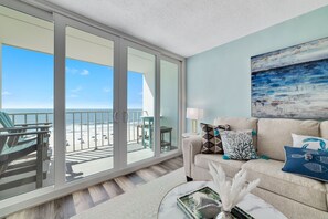 Uma sala de estar ampla com uma grande porta de vidro deslizante que dá para uma varanda com vista para a praia.