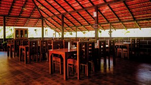 A spacious indoor area with wooden tables and chairs arranged in rows, a red metal roof, and large windows allowing natural light. - Tapasya Home (Virajpet)