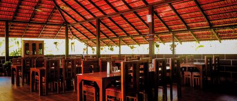 A spacious indoor area with wooden tables and chairs arranged in rows, a red metal roof, and large windows allowing natural light.