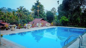 A swimming pool with lounge chairs and tables, surrounded by palm trees and a red-roofed building. - Tapasya Home (Virajpet)