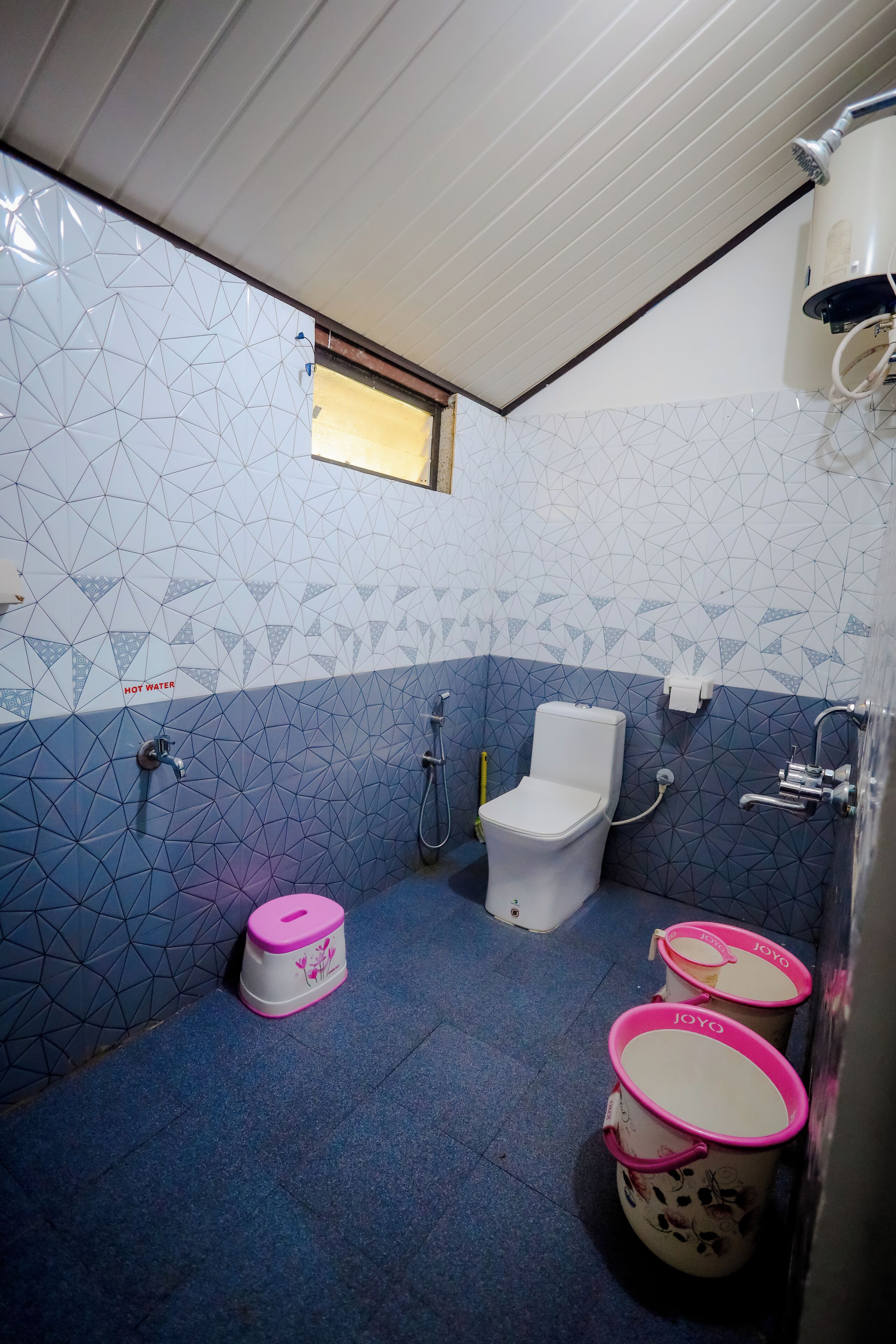 A bathroom with a geometric tile pattern, a toilet, a shower, and three buckets labeled "JOYO".