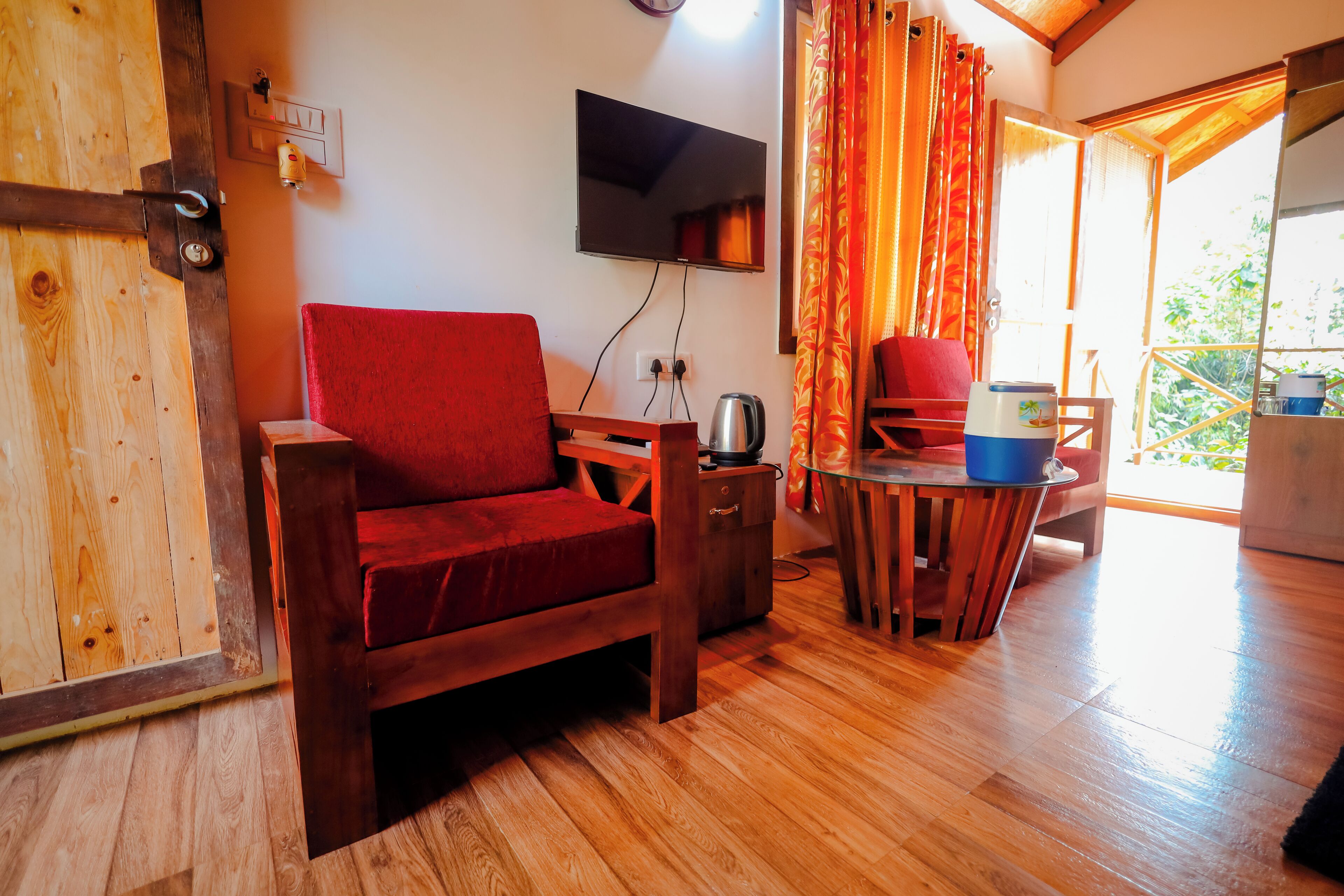 A wooden-floored room with a red sofa, a TV mounted on the wall, and a table with a kettle and a blue container.