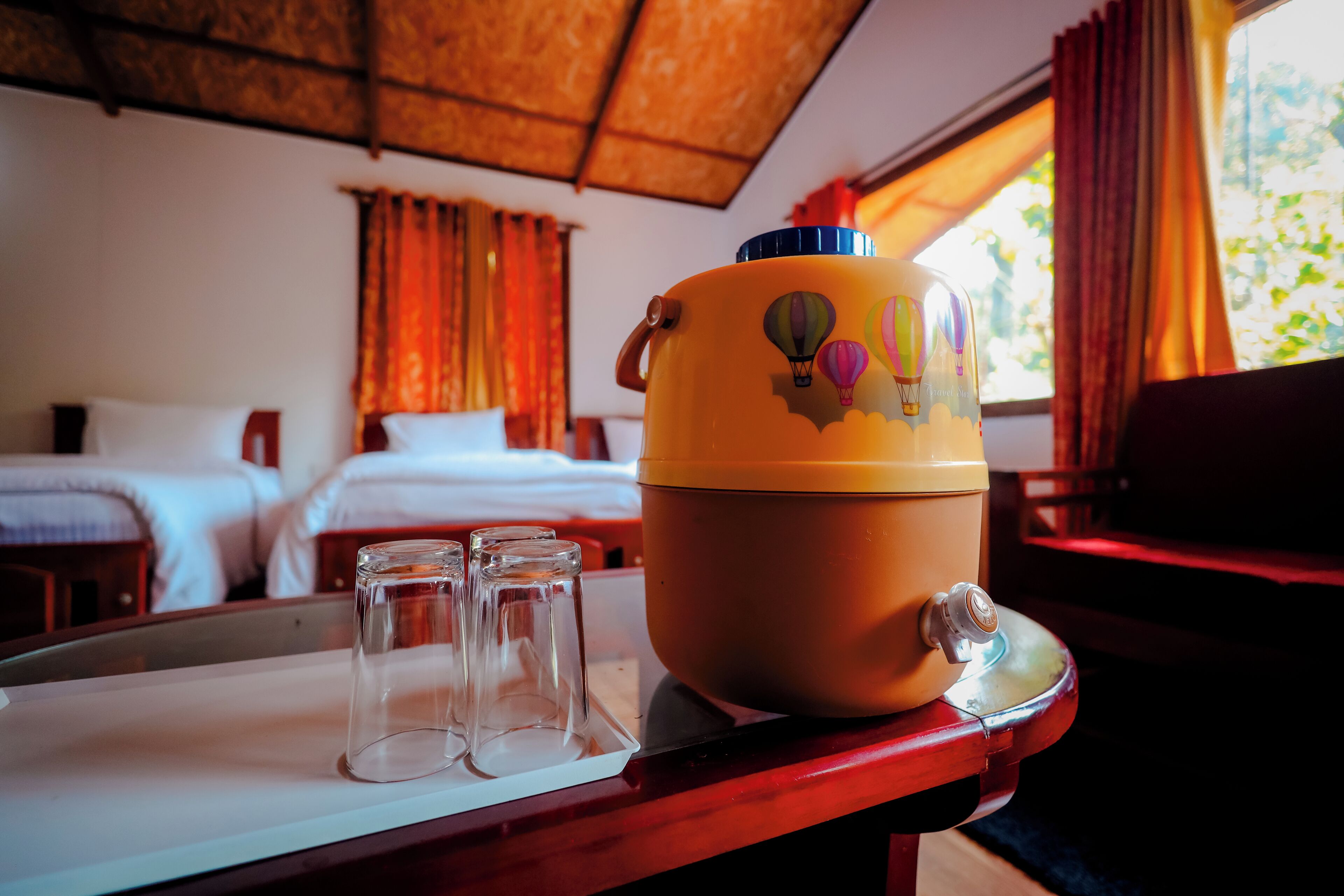 Hotelkamer met een bed, een raam met gordijnen, een tafel met thermoskan en glazen, en een houten plafond.