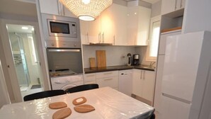 Eine moderne Küche mit weißen Schränken, einem Edelstahlbackofen, einer Mikrowelle und einem Kühlschrank. Ein Esstisch mit weißer Tischdecke und vier Holz Untersetzern.
