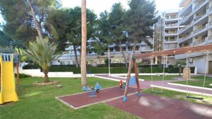 Un terrain de jeux avec des balançoires et un toboggan, entouré de pelouse et d’arbres, avec un bâtiment en arrière-plan.