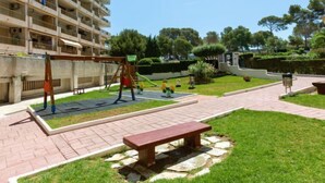 Une aire de jeux avec des balançoires et des toboggans, un banc et une zone dallée avec de l’herbe.