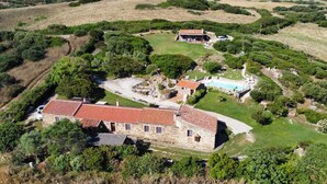 A stone building with a red-tiled roof, surrounded by lush greenery and a swimming pool.