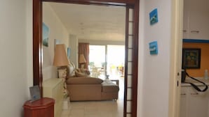 Quarto de hotel com sofá, uma mesinha e uma luminária. Há uma porta dando acesso a outro quarto.